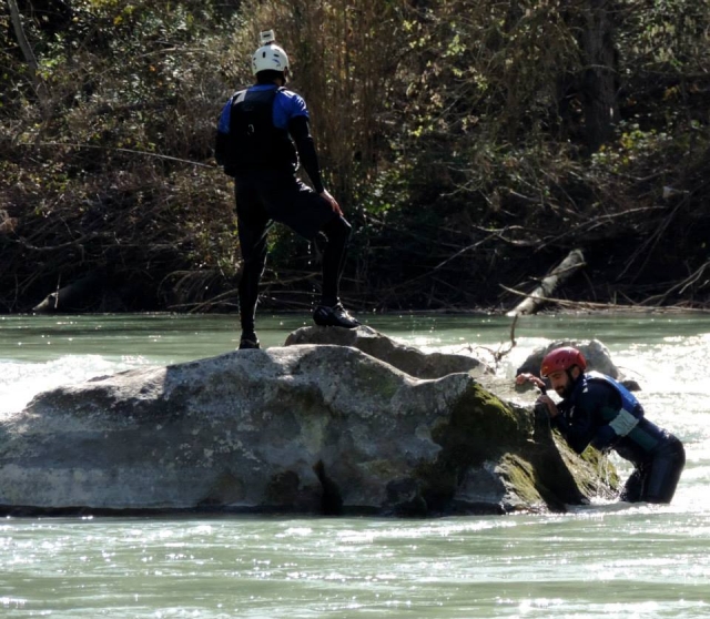  a man climbing a river rock while a man stands on the rock